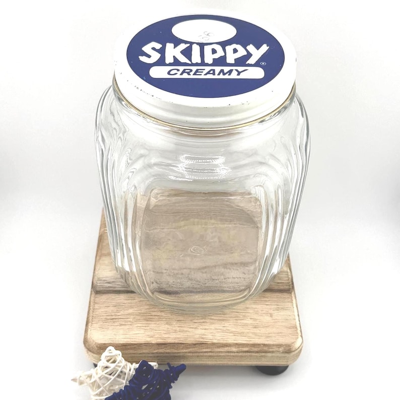 May include: A clear glass jar with a white and blue lid that reads "Skippy Creamy." The jar has a square base and a ridged design. It sits on a small wooden stand with decorative accents.