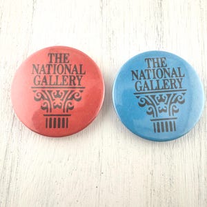 May include: Two round buttons, one red and one blue, each with the text "THE NATIONAL GALLERY" in black. Below the text is a decorative black design. The buttons are likely promotional items or souvenirs.