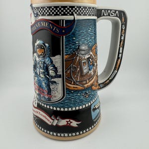 May include: A ceramic beer stein with a black and gold checkered band around the top. The stein features a colorful image of an astronaut on the moon, a lunar module, and a rocket. The text "NASA" is printed on the handle. The text "Achievements" and "Century of Excellence" are printed on the stein.