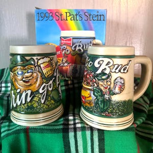 May include: Three ceramic beer steins with green and beige accents, featuring St. Patrick's Day themes. One stein has a leprechaun with sunglasses, another has a leprechaun holding a beer, and the third has the text "Bud" and "1993 St. Pat's Stein" on the box.