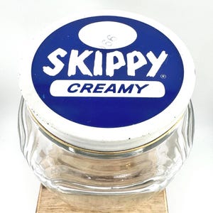 May include: A clear glass jar with a white and blue lid. The lid features the word "SKIPPY" in white letters above the word "CREAMY" in white letters. The jar has a gold-colored rim.