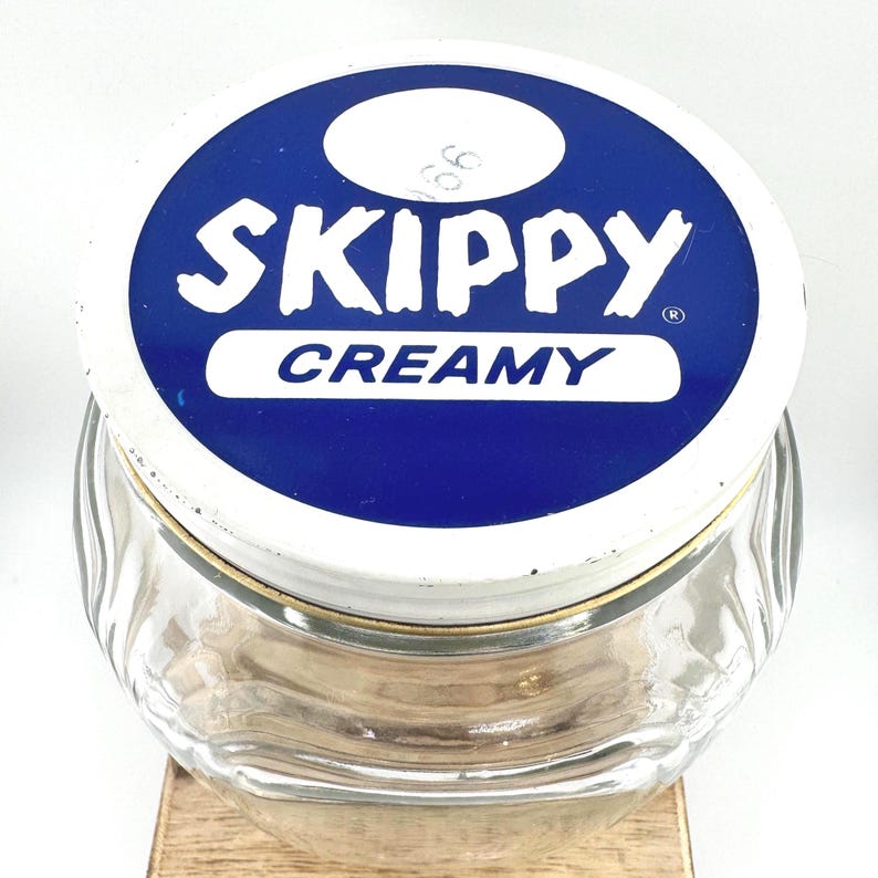 May include: A clear glass jar with a white and blue lid. The lid features the word "SKIPPY" in white letters above the word "CREAMY" in white letters. The jar has a gold-colored rim.