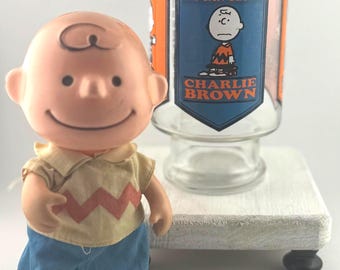 Può includere: Una statuetta di Charlie Brown è accanto a un bicchiere trasparente con il testo "I NEED ALL THE FRIENDS I CAN GET!" e "CHARLIE BROWN". La statuetta indossa una camicia gialla con un motivo a zig-zag rosso e pantaloncini blu. Il bicchiere è su una piattaforma bianca.