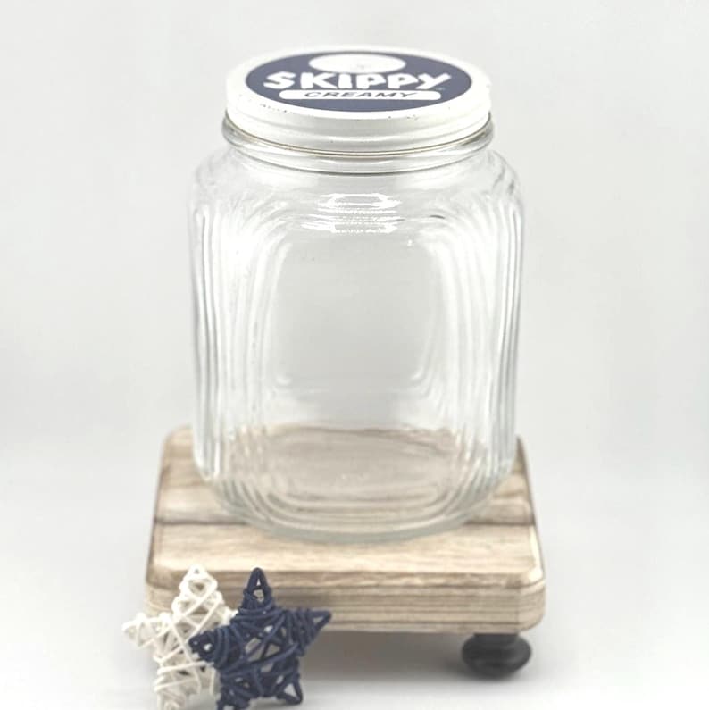 May include: A clear glass jar with a metal lid, labeled "SKIPPY CREAMY" in white and blue. The jar sits on a small wooden stand with decorative stars. The jar has a ribbed design.