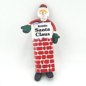Puede incluir: Una figura de Papá Noel flexible. La parte superior de la figura es la cabeza de Papá Noel, con un gorro rojo y barba blanca. El cuerpo es una chimenea de ladrillo rojo con las palabras "Bendable Santa Claus" en la parte delantera. Guantes verdes y zapatos negros completan el look.