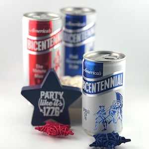 May include: A patriotic display featuring three vintage "America's Bicentennial" beverage cans in red, white, and blue. A navy blue star-shaped decoration with the text "Party like it's 1776" is in front, along with red and blue star-shaped objects.