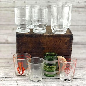 May include: A selection of vintage shot glasses and small measuring cups. Some glasses feature text including "Eastern", "Chef", "Tea", and "Little America Wyoming". The glasses are clear with red and green details, arranged on a wooden box.