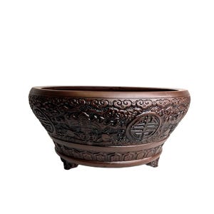 May include: A brown, intricately carved ceramic bowl with a wide, oval shape. The bowl features detailed relief patterns of traditional Asian motifs, including symbols and dragons. It rests on three small feet.