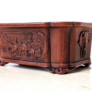 May include: A rectangular terracotta bonsai pot with a raised rim and four feet. The pot is decorated with a relief design of elephants in a forest setting.