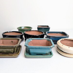 May include: Assortment of eight ceramic bonsai pots in various shapes and colours, including green, blue, black, and white. The pots are arranged on a white surface.
