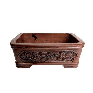 May include: A square terracotta bonsai pot with a decorative floral design. The pot has a brown glaze and is approximately 25 cm wide.