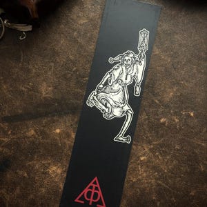 May include: Black bookmark with a white illustration of a jester holding a lantern. The jester has a skeletal appearance and is in motion. A red triangle with a symbol is at the bottom. The bookmark is rectangular.
