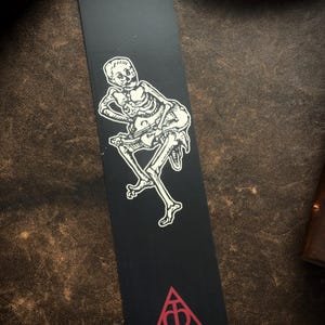 May include: Black bookmark with a white illustration of a skeleton playing a stringed instrument. A red triangle with a symbol is at the bottom. The bookmark is rectangular and has a matte finish.