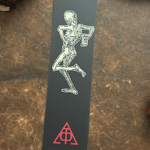 May include: Black bookmark with a white illustration of a skeleton carrying a mug. A red symbol is at the bottom. The bookmark is rectangular and made of card. The overall theme is gothic and macabre.