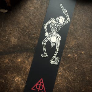 May include: Black bookmark with a white illustration of a skeleton playing a drum and holding a drumstick. The bottom of the bookmark has a red triangle with a symbol inside. The bookmark is rectangular.