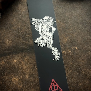 May include: Black bookmark with a white illustration of a skeleton playing a musical instrument. A red triangle with a symbol is at the bottom. The bookmark is rectangular and made of a matte material.