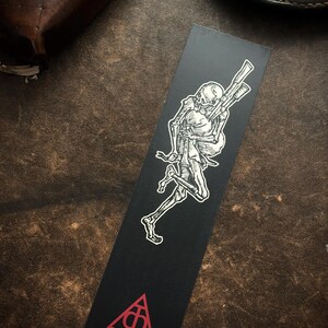 May include: Black bookmark with a white illustration of a skeleton carrying rifles. A red triangle with a symbol is at the base. The bookmark is rectangular, possibly made of card or paper.