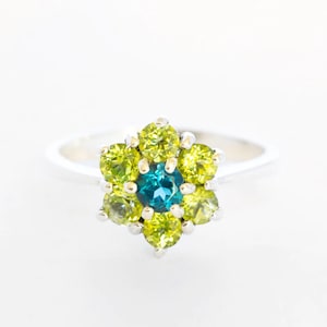 Op de afbeelding: Een zilveren ring met een cluster van groene en blauwe edelstenen, gezet in de vorm van een bloem.