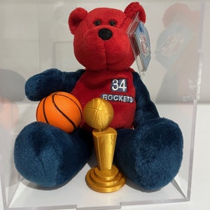 May include: A red and blue plush teddy bear wearing a red jersey with the number 34 and the word "ROCKETS" on it. The bear is holding a basketball and a gold trophy.