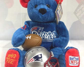 May include: A blue plush teddy bear with red ears and feet, wearing a Patriots logo on its chest. The bear holds a brown football and sits behind a silver football helmet with a red facemask. The feet feature embroidered years and an NFL logo.