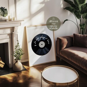 May include: A framed print featuring a black vinyl record design with the text "if it's nice play it twice" in white script. The print is displayed in a room with a neutral color palette, a brown sofa, and a white coffee table.