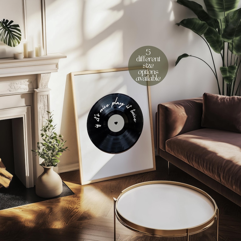 May include: A framed print featuring a black vinyl record design with the text "if it's nice play it twice" in white script. The print is displayed in a room with a neutral color palette, a brown sofa, and a white coffee table.
