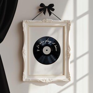 May include: A framed print featuring a black vinyl record design. The record has white text that reads "if it's nice play it twice" and a small heart. The frame is white with ornate detailing, and a black bow hangs above.