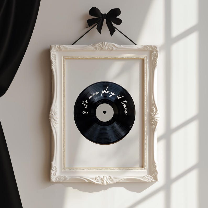 May include: A framed print featuring a black vinyl record design. The record has white text that reads "if it's nice play it twice" and a small heart. The frame is white with ornate detailing, and a black bow hangs above.