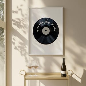 May include: A framed print featuring a black vinyl record with the text "if it's nice play it twice" and a small heart. The print is in a white frame. A gold bar cart holds wine glasses and a wine bottle.