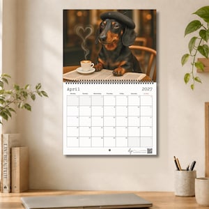 May include: A wall calendar for April 2027 featuring a black and tan dachshund wearing a dark beret, sitting at a table with a newspaper and a cup of coffee with heart-shaped steam. The calendar grid displays the month, year, and days of the week.