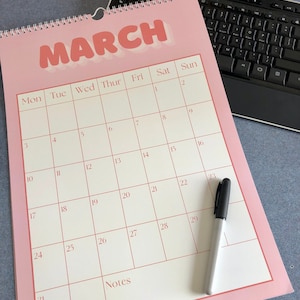 May include: A pink monthly calendar with the month of March highlighted in bold, pink letters. The calendar has a white background with black numbers and a black marker on the right side.