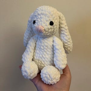 May include: A white crocheted bunny rabbit plush toy with black eyes and a pink nose. The bunny is sitting on a hand.