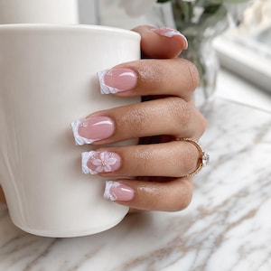 Può includere: Una mano con unghie curate, che tiene una tazza bianca. Le unghie presentano una base rosa con accenti bianchi simili a pizzo e piccoli disegni floreali. Un anello d'oro con un diamante è sull'anulare.