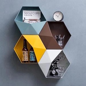 May include: A geometric wall shelf unit with six hexagonal compartments in various colours, including blue, brown, yellow, and white. The shelves are filled with items such as books, bottles, and a vintage projector.