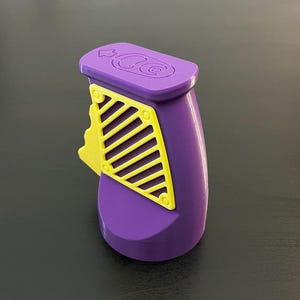 K&ouml;nnte beinhalten: Eine lila-gelbe Spielzeugpistole mit Abzug und einem gelben gitterartigen Design an der Seite. Die Oberseite der Pistole hat ein geschwungenes Design. Die Spielzeugpistole ist aus Kunststoff und steht auf einer dunklen Oberfl&auml;che.