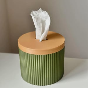 May include: A cylindrical tissue box with a light brown lid and a green, vertically ribbed body. White tissues are pulled from the top. The tissue box sits on a white surface.