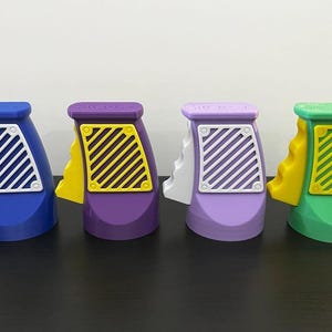 K&ouml;nnte beinhalten: Vier bunte Spielzeugpistolen in Blau, Lila, Lavendel und Gr&uuml;n. Jede hat einen wei&szlig;en und gelben Akzent mit einem diagonal gestreiften Design an der Seite. Die Spielzeuge sind in einer Reihe auf einer dunklen Oberfl&auml;che angeordnet.