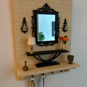 May include: A decorative wall-mounted key holder with a small mirror, shelf, and key hooks. The black mirror has an ornate frame, and the shelf holds a lamp, vase, and flowers. The background has a brick-like pattern.