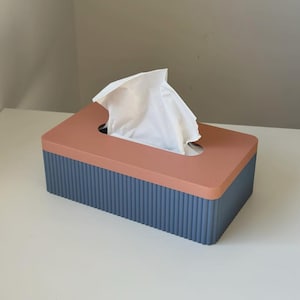 May include: A rectangular tissue box with a coral-coloured top and a blue ribbed base. White tissues are visible, emerging from the opening. The box is made of a solid material and sits on a white surface.