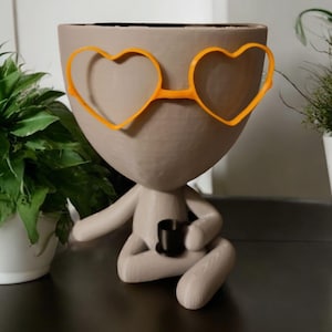 May include: A whimsical, light gray planter shaped like a person, seated with legs crossed. It features orange heart-shaped glasses and holds a small black cup. The planter is designed to hold small plants and adds a touch of personality to any space.
