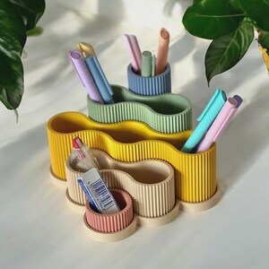 May include: A set of colourful desk organisers in yellow, green, and beige, each with a wavy design and cylindrical pen holders. The organisers hold pens and pencils in various pastel colours. A small pink container holds an eraser and other small items.