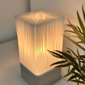 Peut inclure: Une lampe de table rectangulaire avec un abat-jour blanc texturé et une base grise. La lampe est allumée, dégageant une lueur chaleureuse. Une plante verte est visible en arrière-plan.