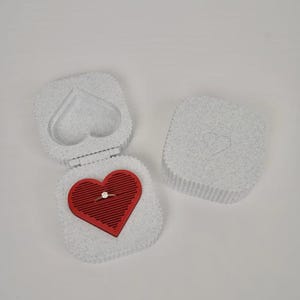 May include: A white, textured ring box with a red heart-shaped insert. The box is open, revealing a gold ring with a diamond. The lid has a heart-shaped cutout. A separate, closed box is also visible.