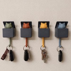 May include: Four wall-mounted key hooks, each featuring a dark grey box with a pop-up cat figure in olive green, terracotta, yellow, and blue. Each hook extends from the bottom of the box, holding a metal keyring with various keys attached.