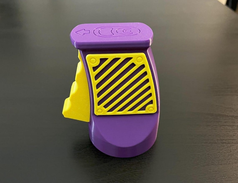 K&ouml;nnte beinhalten: Ein lila-gelbes Kunststoffger&auml;t mit einer diagonalen gelb-schwarz gestreiften Platte. Die Oberseite hat einen Pfeil und ein Kreissymbol. Das Ger&auml;t hat einen gelben Hebel an der Seite.