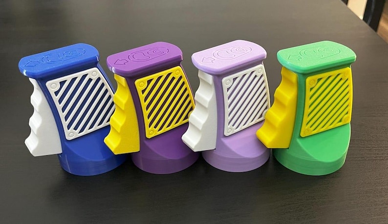 K&ouml;nnte beinhalten: Vier farbenfrohe, 3D-gedruckte Objekte in Blau, Lila, Lavendel und Gr&uuml;n. Jedes hat einen wei&szlig;en Griff, einen gelben Akzent und ein wei&szlig;es oder gelbes Gitter. Die Oberseiten haben Pfeile und kreisf&ouml;rmige Designs. Die Objekte stehen auf einer dunklen Oberfl&auml;che.