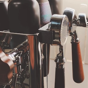 May include: A close-up of a silver espresso machine with two chrome portafilters and four black ceramic mugs. The machine has a wooden handle and a silver lever.
