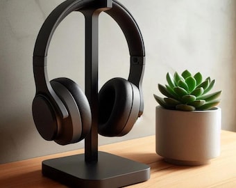 Puede incluir: Auriculares negros con un soporte negro sobre una superficie de madera. Una pequeña planta suculenta en maceta se encuentra a la derecha del soporte.