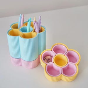 Peut inclure: Un ensemble d'organisateurs de bureau en forme de fleur aux couleurs pastel. L'ensemble comprend un porte-stylos avec des sections bleues et roses, et un plateau séparé avec des compartiments jaunes et roses. Des stylos et des trombones sont visibles.
