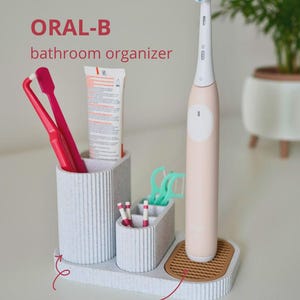 May include: A light grey bathroom organiser with the text "ORAL-B bathroom organiser". It holds a pink electric toothbrush, two red toothbrushes, toothpaste, and dental floss. Includes a practical drip tray.